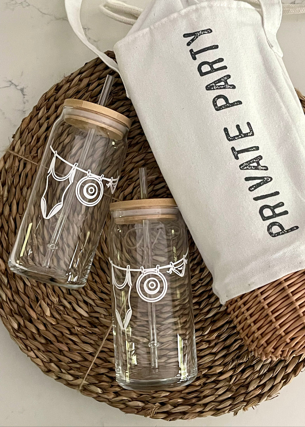 Two glass tumblers with wooden lids and straws, accompanied by a white drawstring bag labeled 'PRIVATE PARTY' on a woven mat.